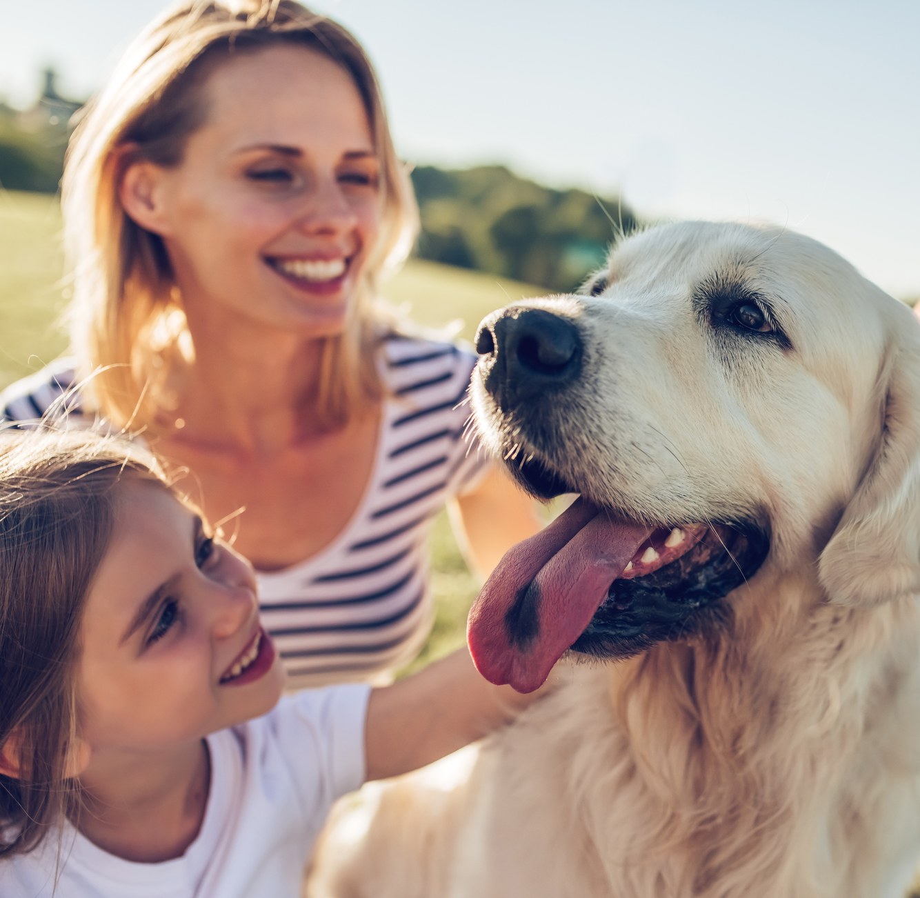 A woman and a young girl smiling beside a golden retriever outdoors
